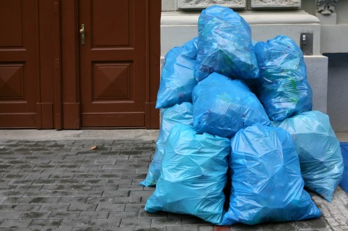 A group of several large blue rubbish bags made of plastic, filled with waste, are stacked on a paved surface outside a building. The bags are situated near a brown wooden door and a grey utility box, with some bags leaning against each other and showing creases and folds in the material. The scene is outdoors, on a grey brick pavement, with a portion of a white wall or window frame visible in the background, indicating a residential or commercial property. The image likely relates to rubbish removal services, such as those provided by Wood Green House Clearance, and depicts a typical collection of waste awaiting collection or disposal in an urban setting within the area covered by the service.