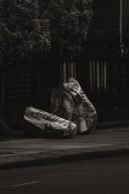 Two large black bagged waste bundles are positioned on the edge of a paved street, leaning slightly against a black metal fence with vertical bars. The bags appear to contain mixed rubbish, with some